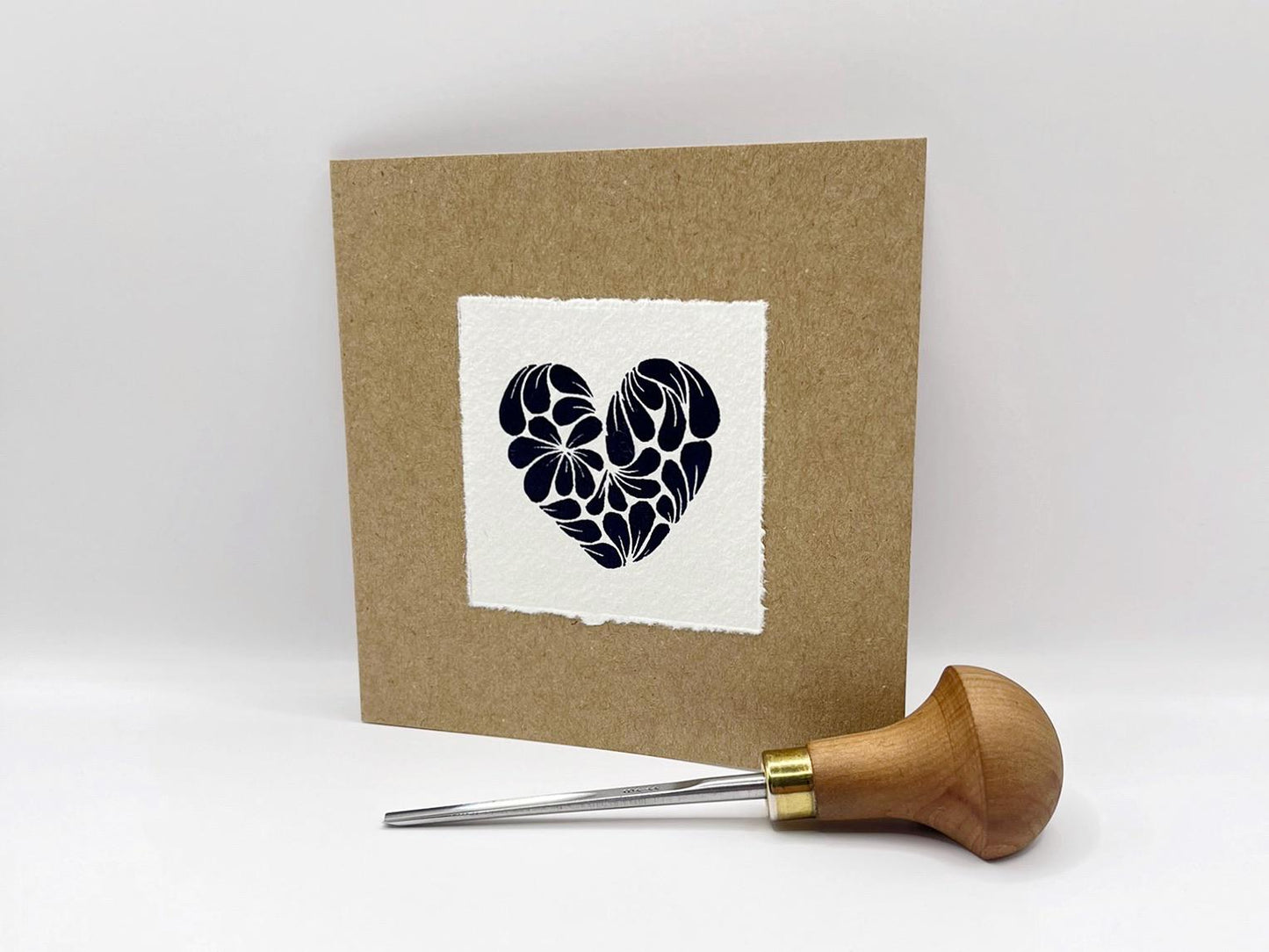 Kraft card with original lino printed blue heart design on white paper stood against a white background. Photo includes lino cutting tool.