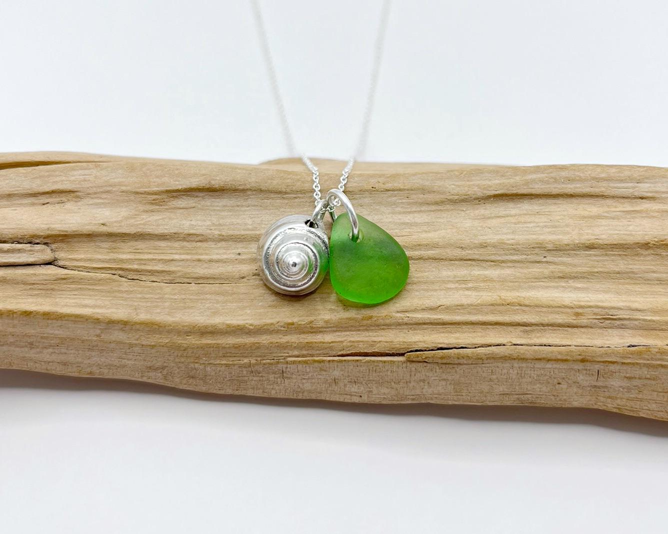Necklace with a silver shell and green sea glass laid on driftwood against a white background
