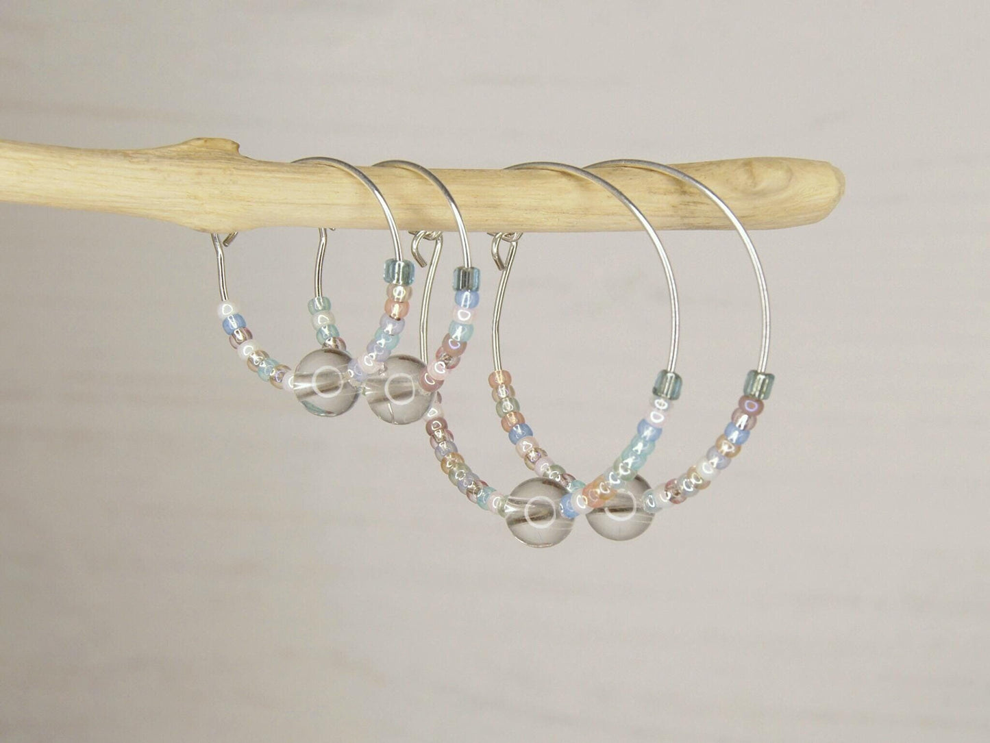 Two pairs of pastel seed bead and clear crystal hoop earrings hanging on driftwood against a pale background