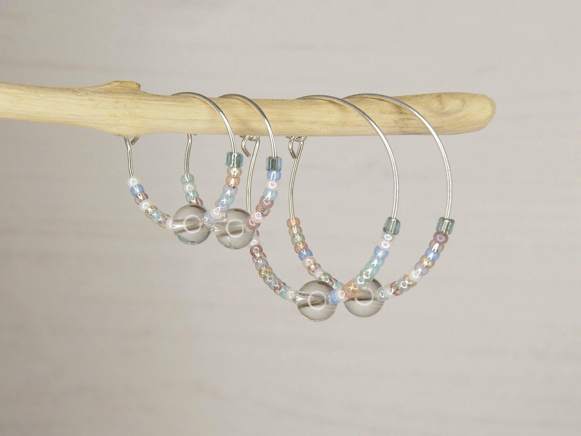 Two pairs of pastel seed bead and clear crystal hoop earrings hanging on driftwood against a pale background