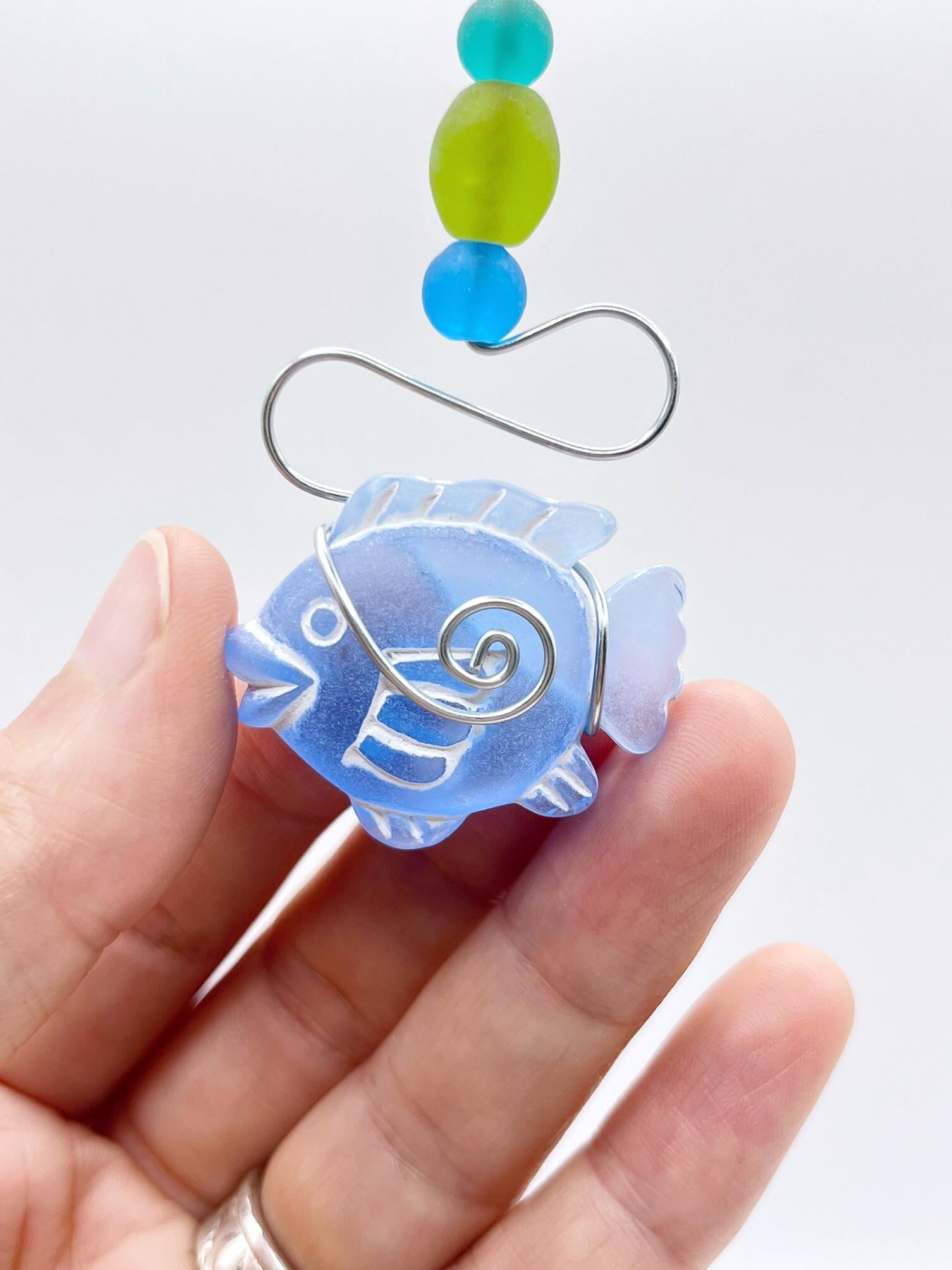 Decorative hanging ornament with blue fish, wire and colourful beads held in a hand against a white background