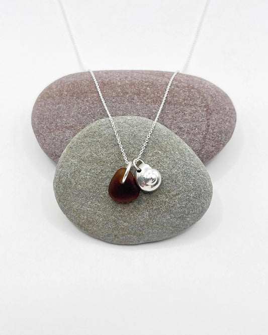 Necklace with burnt orange sea glass and a silver shell pendant, laid across two pebbles on a white surface