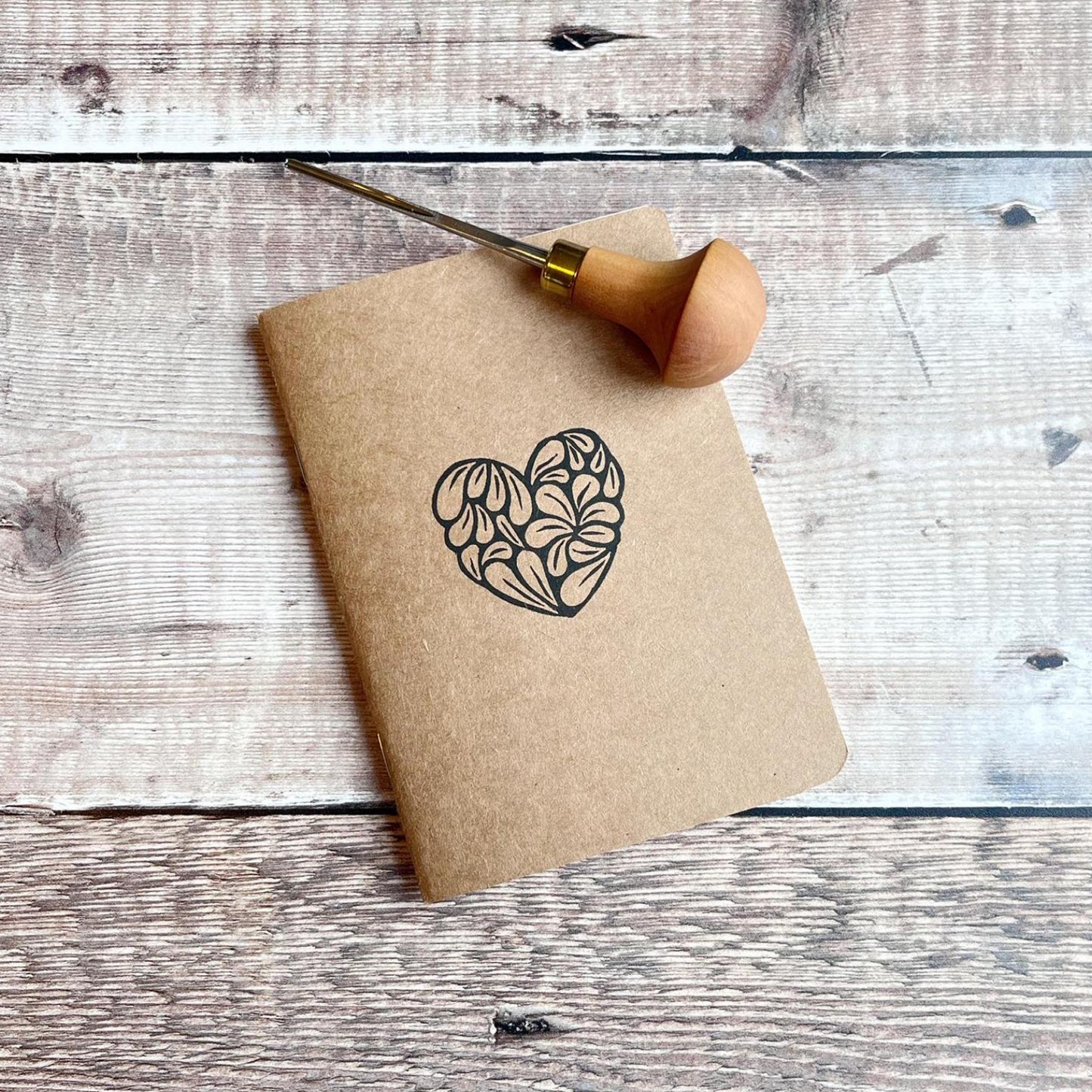 Kraft paper notebook with an original lino print heart design laid on a wooden surface 