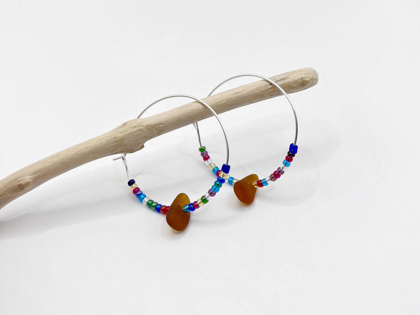 Rainbow seed bead and burnt orange sea glass hoop earrings hanging on driftwood against a white background