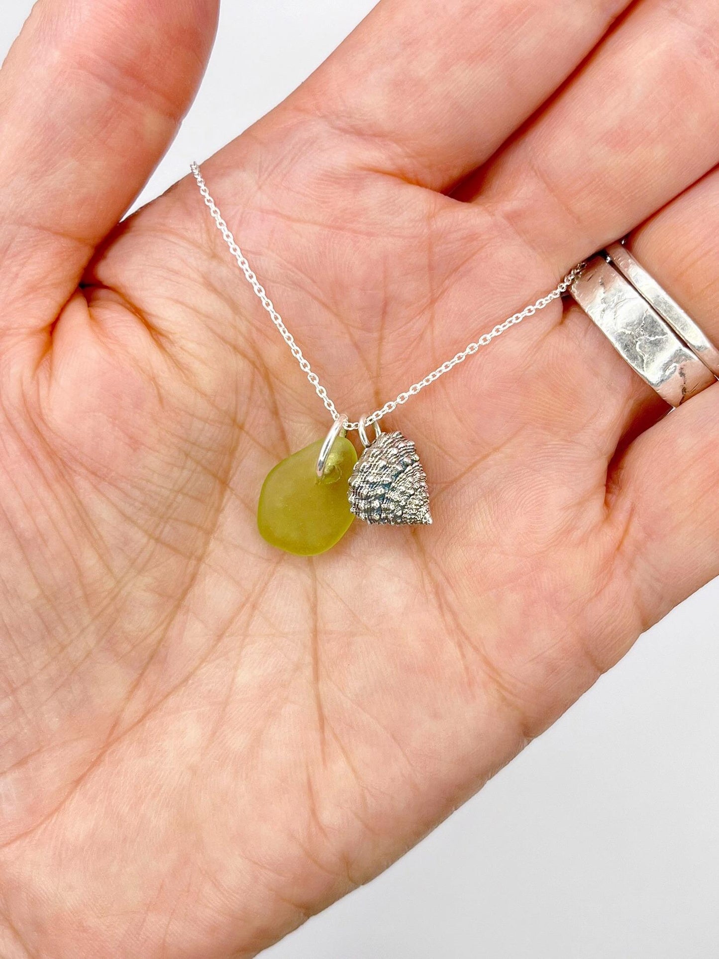 Hand holding a necklace with lime green sea glass and a silver shell charm against a white background