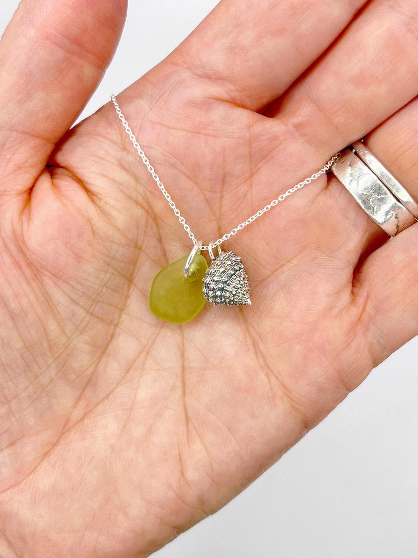 Hand holding a necklace with lime green sea glass and a silver shell charm against a white background