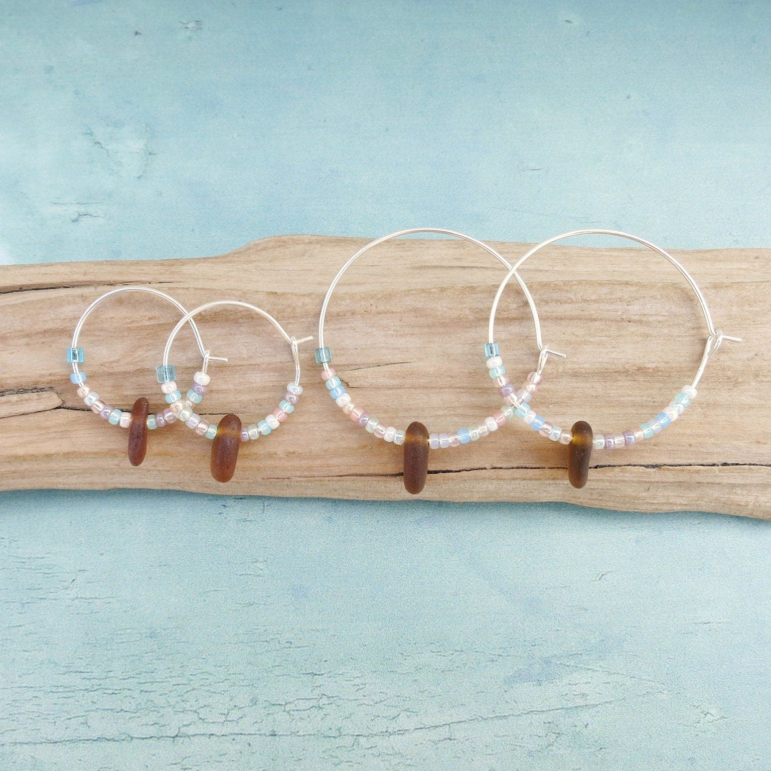 Two pairs of silver hoop earrings with pastel seed beads and burnt orange sea glass, laid on a piece of driftwood with a light blue background