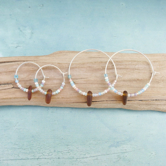 Two pairs of silver hoop earrings with pastel seed beads and burnt orange sea glass, laid on a piece of driftwood with a light blue background