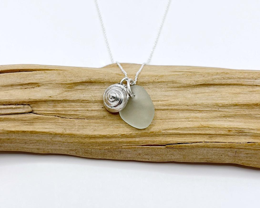 Necklace with a silver shell and seaglass pendant laid on driftwood against a white background