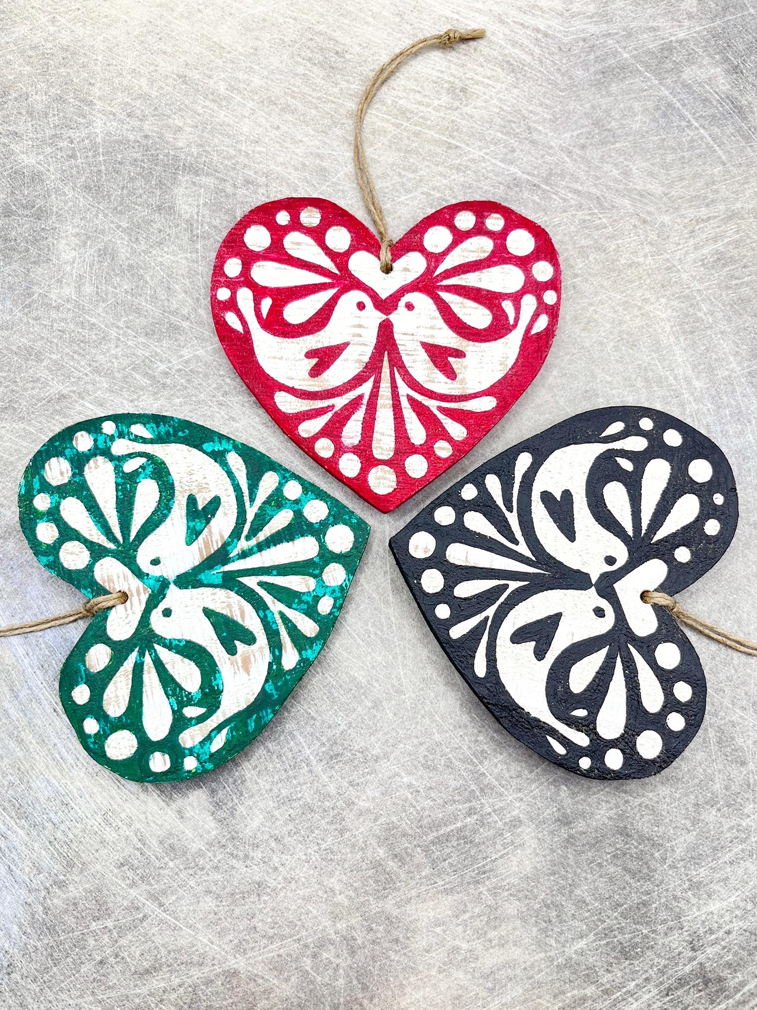 Three heart-shaped wooden hanging decorations with folk style love bird print   in red, green, and black laid on a textured surface.