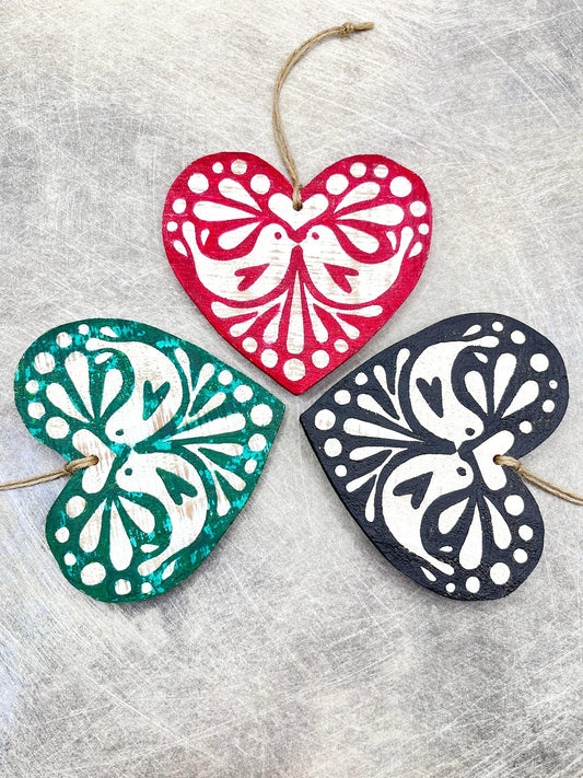 Three heart-shaped wooden hanging decorations with folk style love bird print   in red, green, and black laid on a textured surface.
