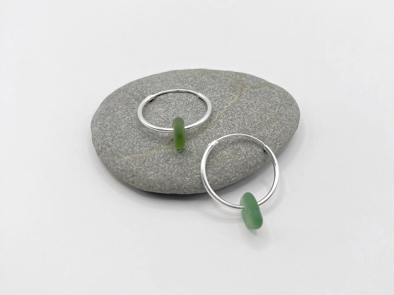 Silver hoop earrings with soft green sea glass laid on a grey pebble against a white background