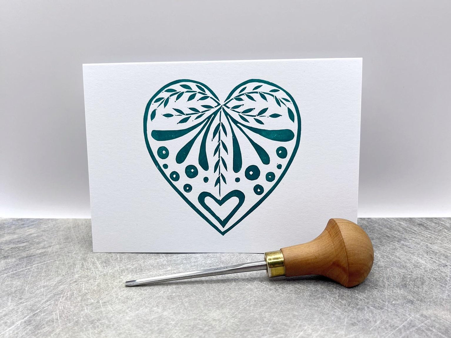 White card with an original lino printed green heart design laid on a textured surface. Photo includes lino cutting tool.