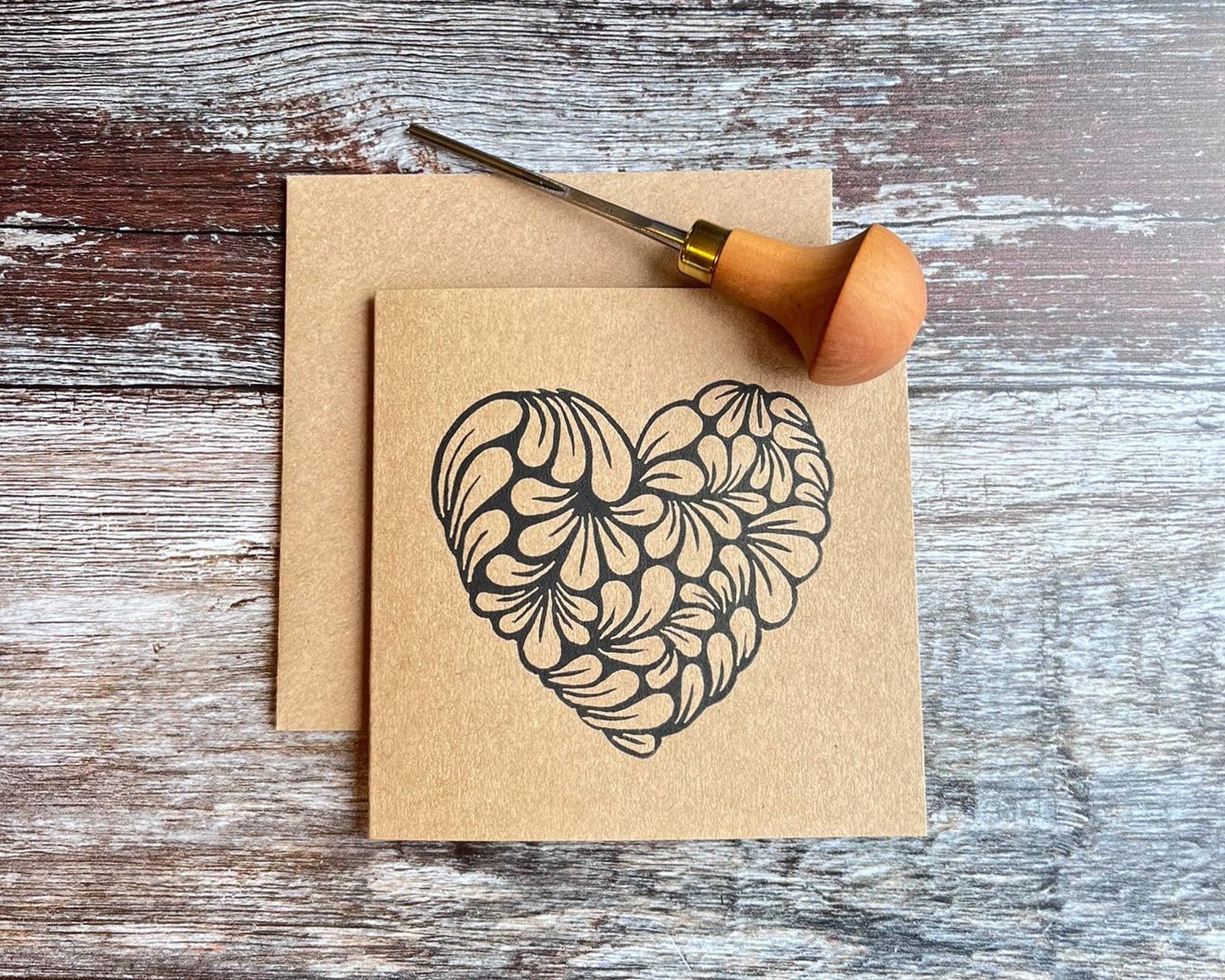 Kraft card with an original lino printed heart laid on a wooden surface. Photo includes lino cutting tool used.