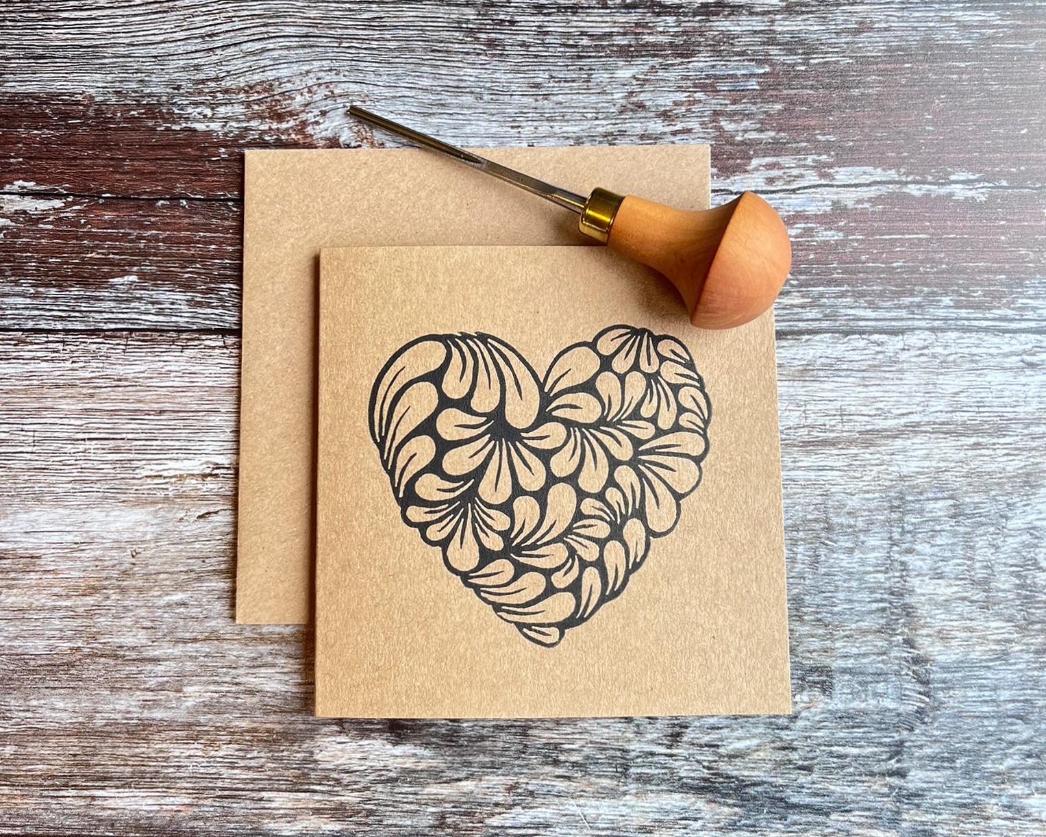 Kraft card with an original lino printed heart laid on a wooden surface. Photo includes lino cutting tool used.