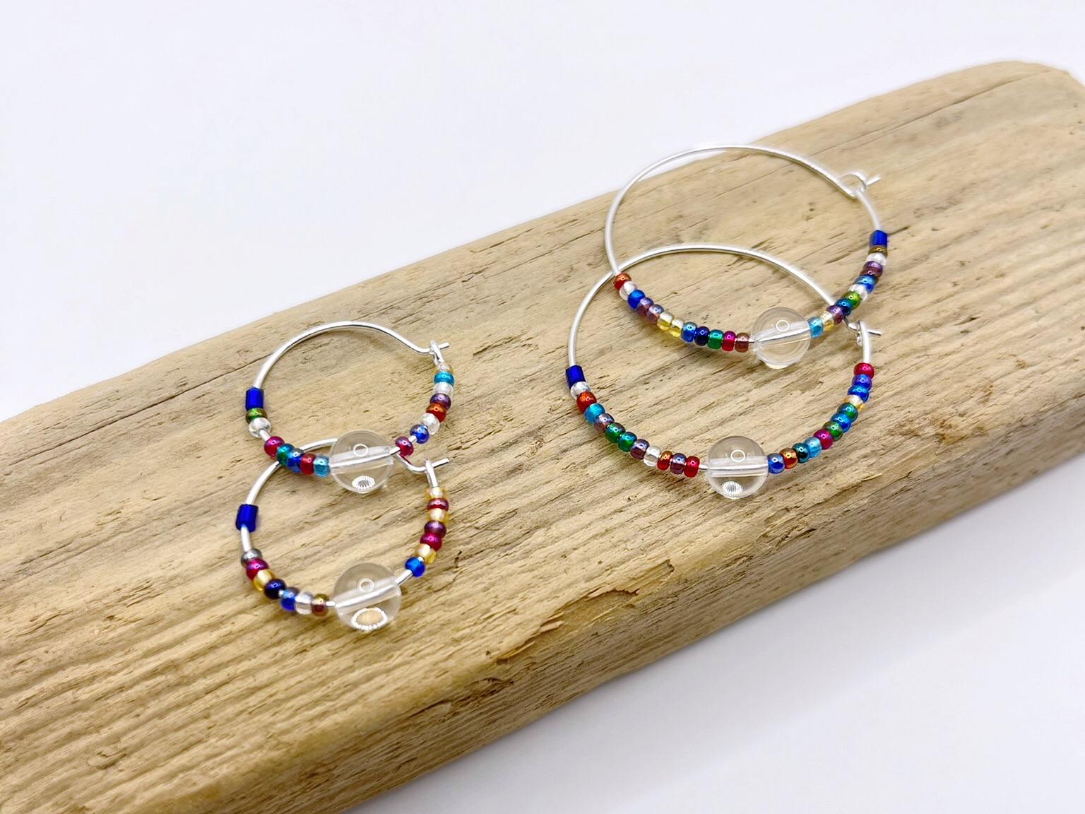 Two pairs of rainbow seed bead and crystal hoop earrings laid on driftwood against a white background.