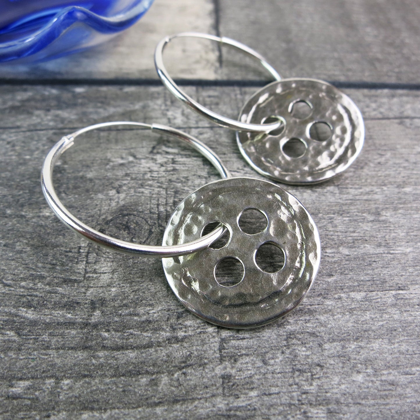 Close up of silver hoop earrings with large round hammered silver buttons laid on a wooden surface.