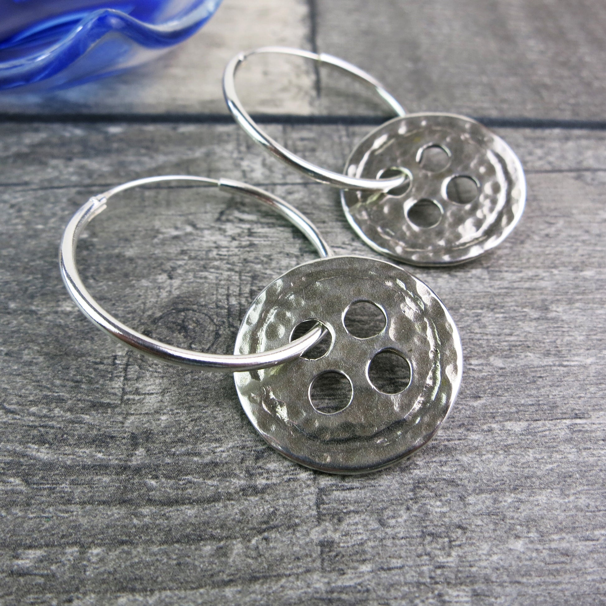 Close up of silver hoop earrings with large round hammered silver buttons laid on a wooden surface.