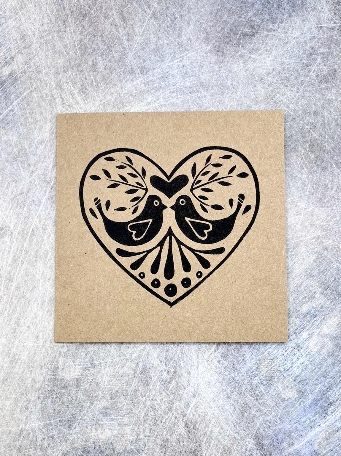Kraft card with an original lino print heart featuring two birds laid on a textured grey background