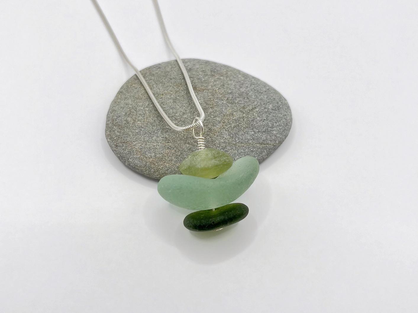 Necklace with green sea glass and Jade pendant sat against a grey pebble on a white background