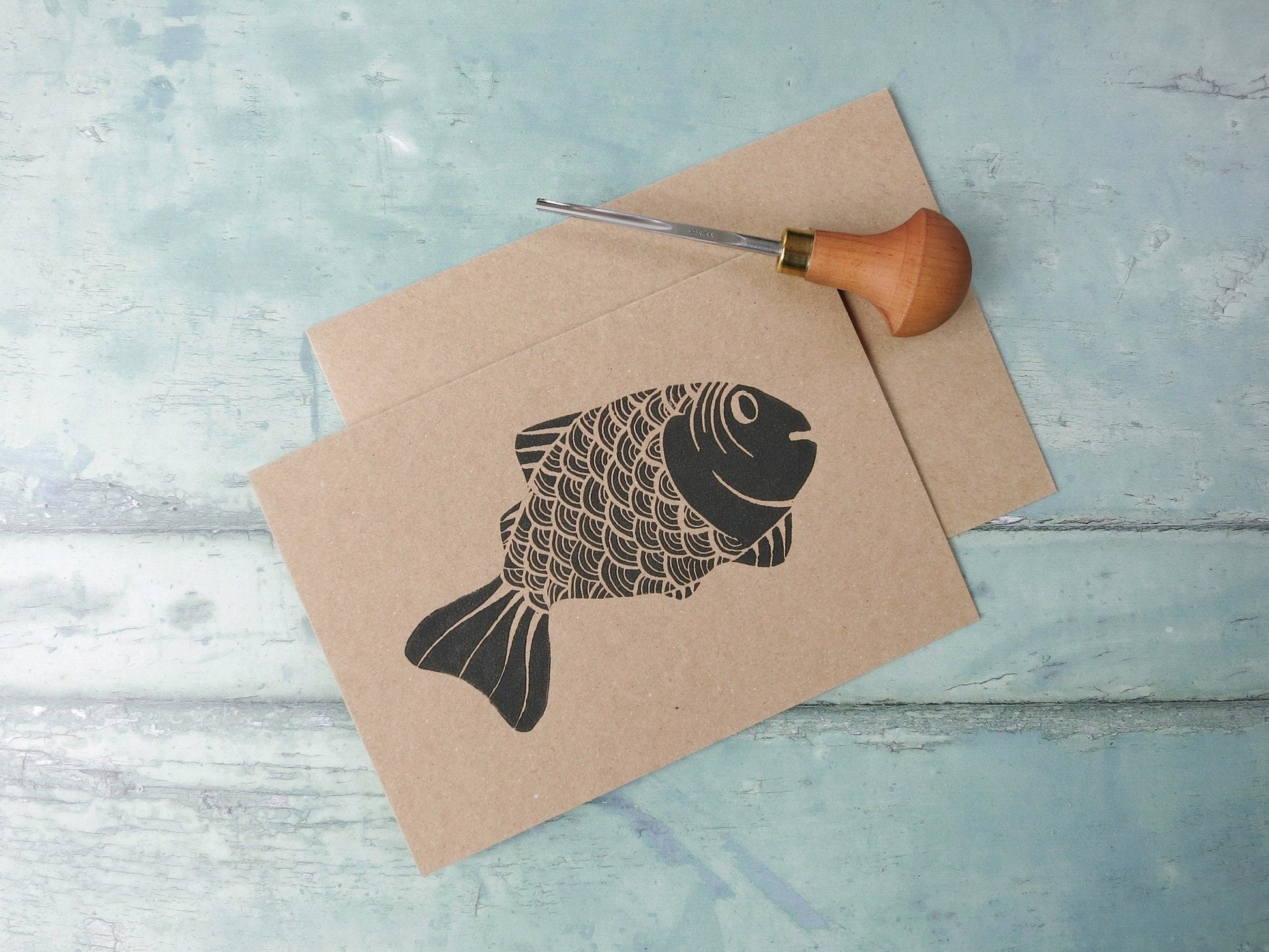 Kraft paper card with original lino printed fish laid on a textured surface. Photo includes lino cutting tool
