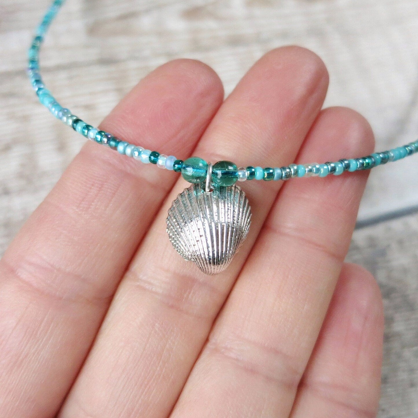 Turquoise beaded necklace with a silver shell charm held in a hand against a neutral background