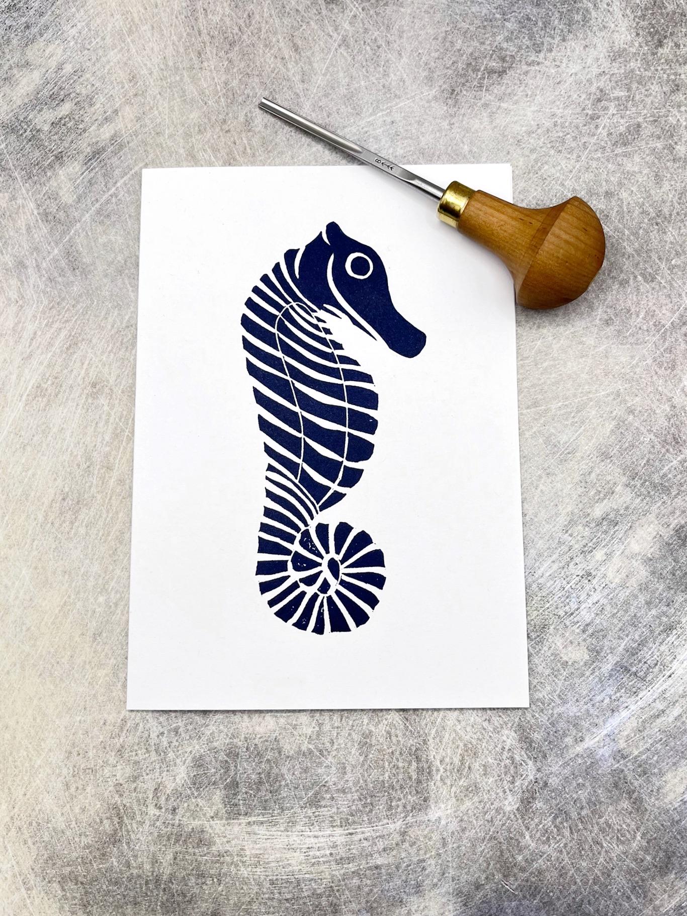 Original lino print of a navy blue seahorse design on a white card, laid on a textured background. Photo includes lino cutting tool.