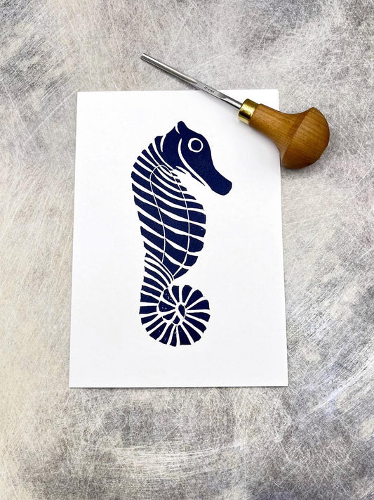 Original lino print of a navy blue seahorse design on a white card, laid on a textured background. Photo includes lino cutting tool.