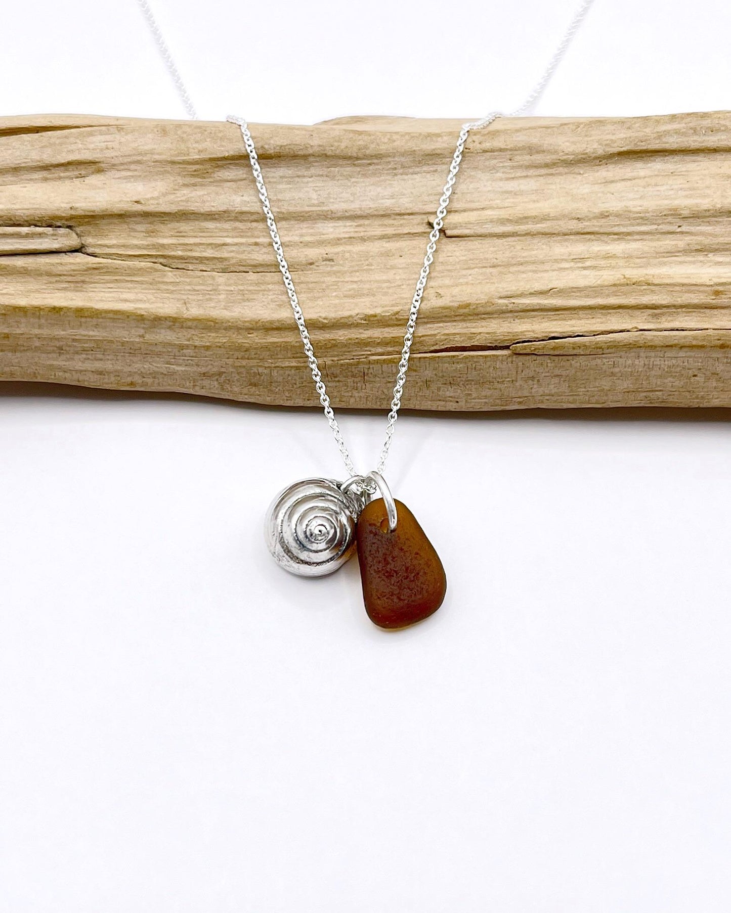 Silver necklace with burnt orange sea glass and a silver shell pendant laid in front of a piece of driftwood against a white background