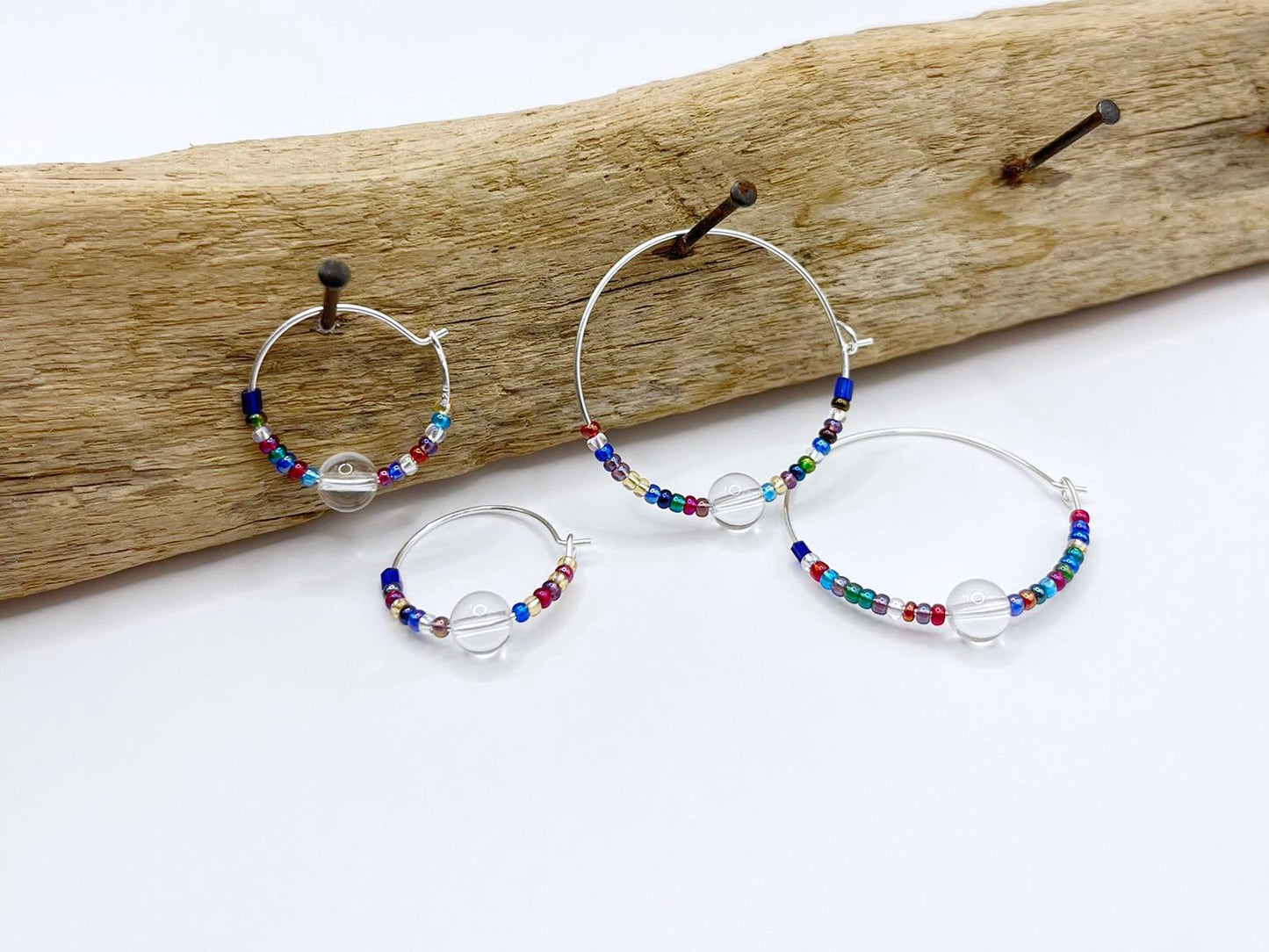 Rainbow seed bead and crystal hoops earrings hanging on driftwood against a white background.