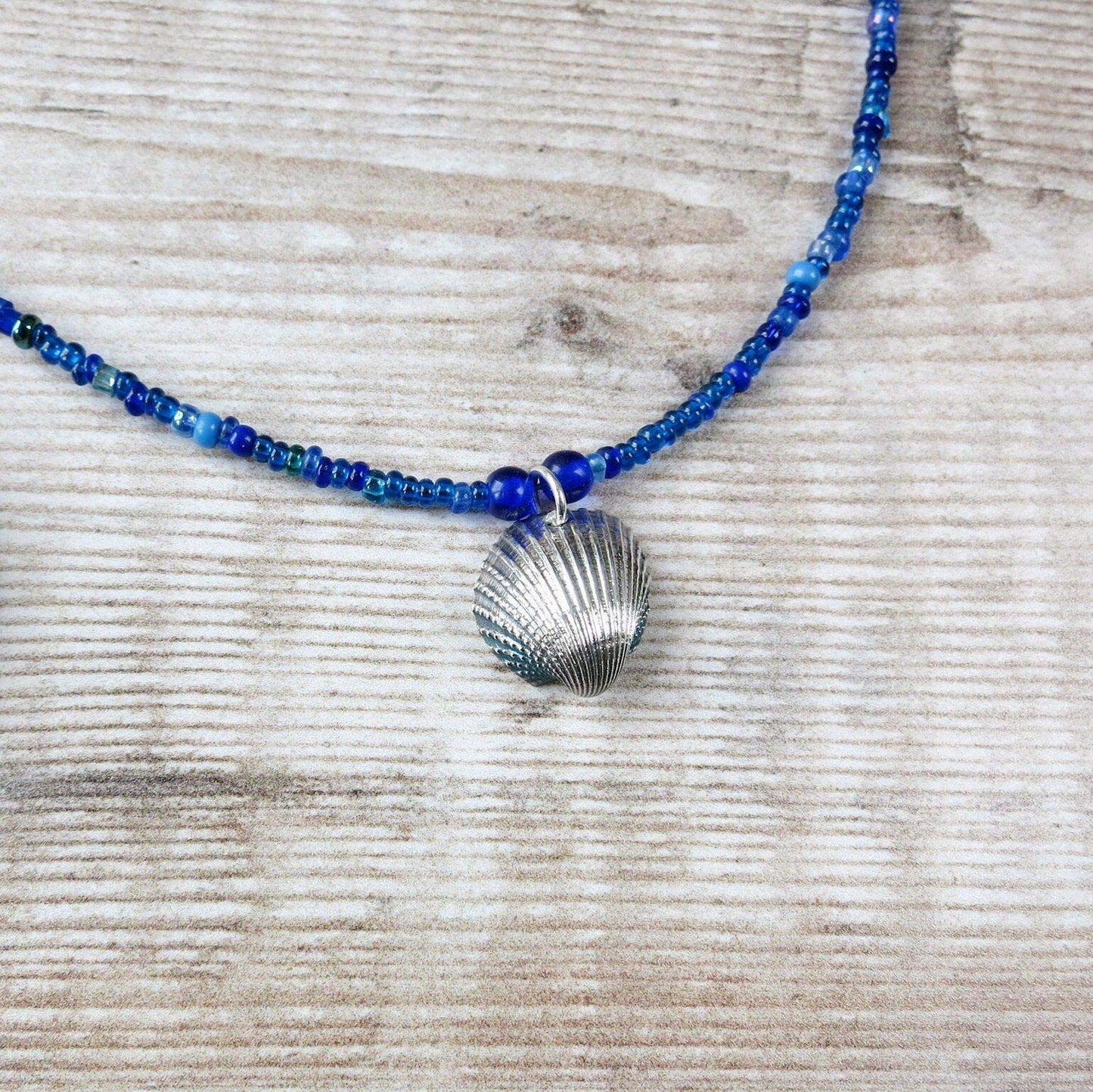 Blue seed bead necklace with silver coated shell pendant laid on a textured surface