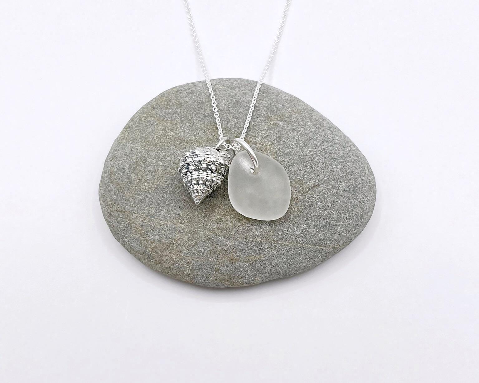 Silver necklace with White Sea glass and silver shell pendant laid on a grey pebble against a white background