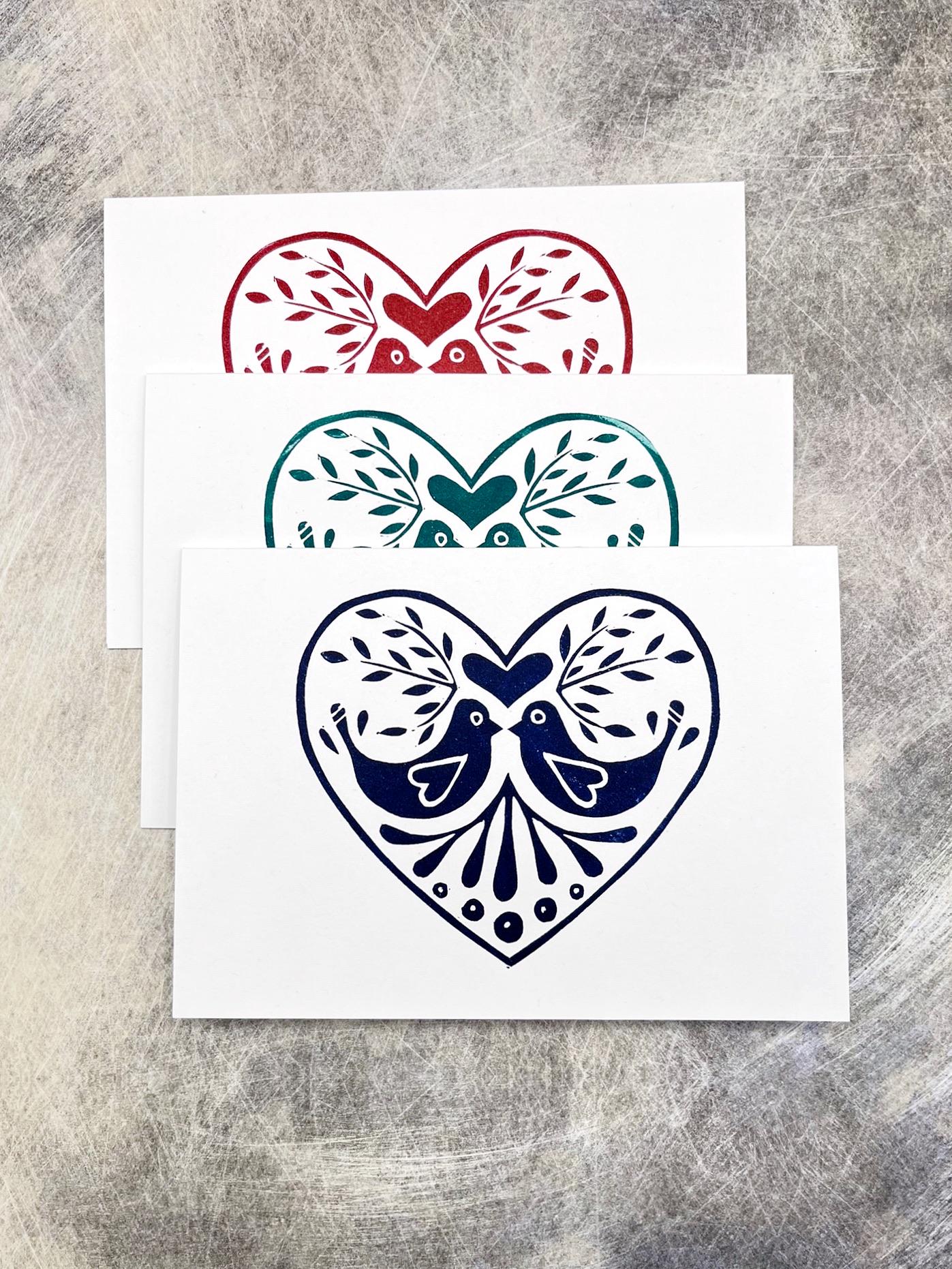 Three original lino printed heart design cards in red, blue and green, laid on a textured surface