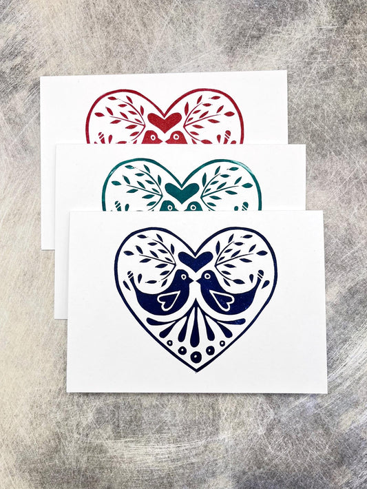 Three original lino printed heart design cards in red, blue and green, laid on a textured surface