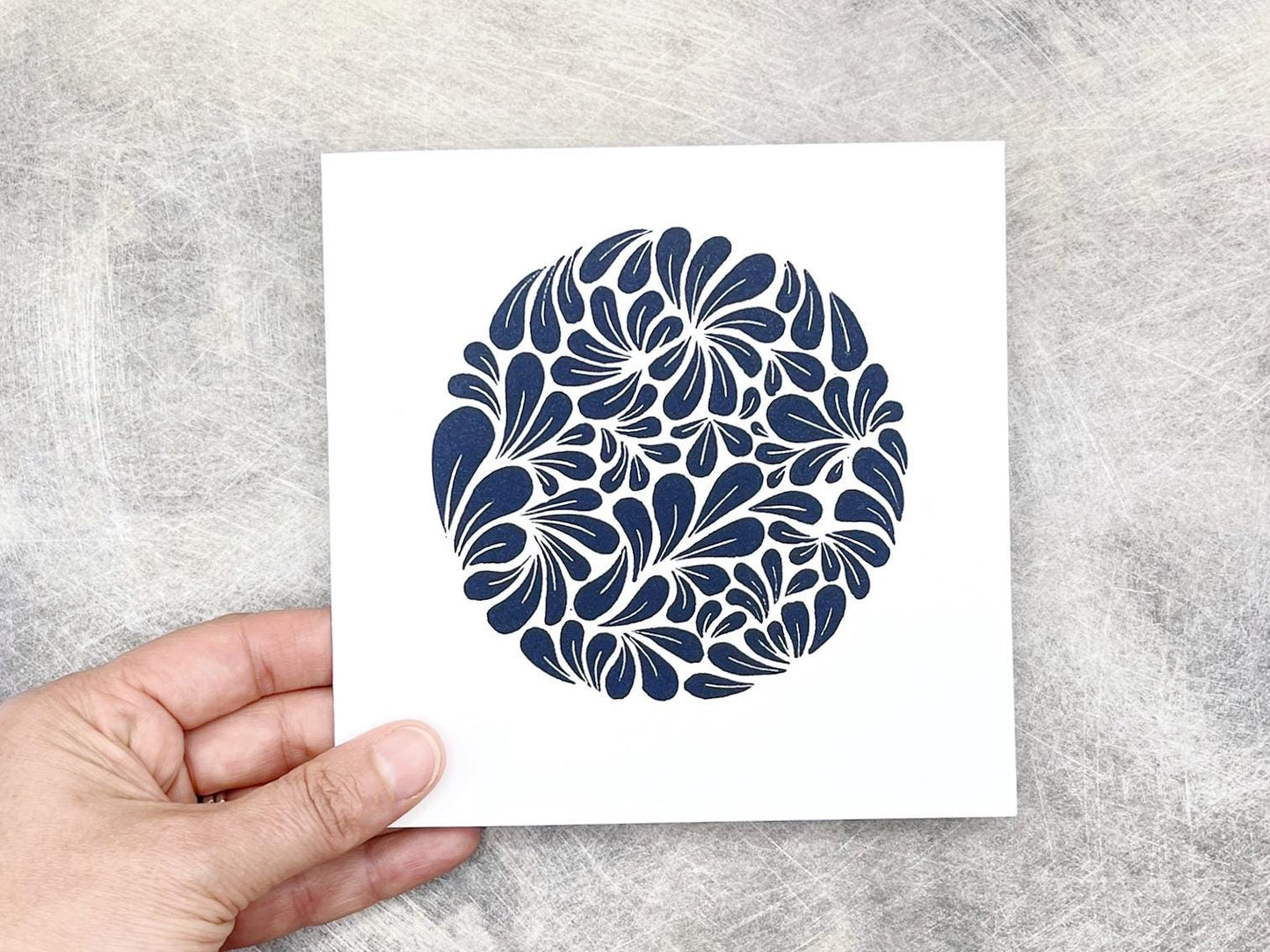 Hand holding white card with navy blue original lino printed mussel shell pattern against a textured background 