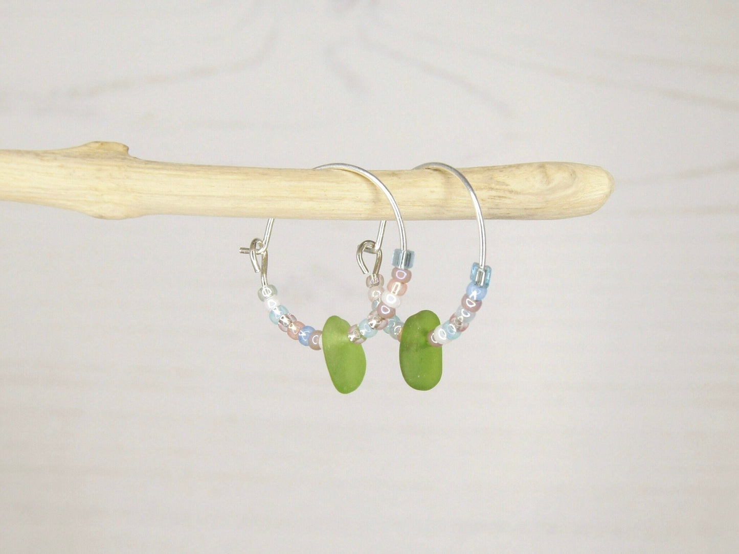 Hoop earrings with pastel seed beads and green sea glass hanging on driftwood against a light background