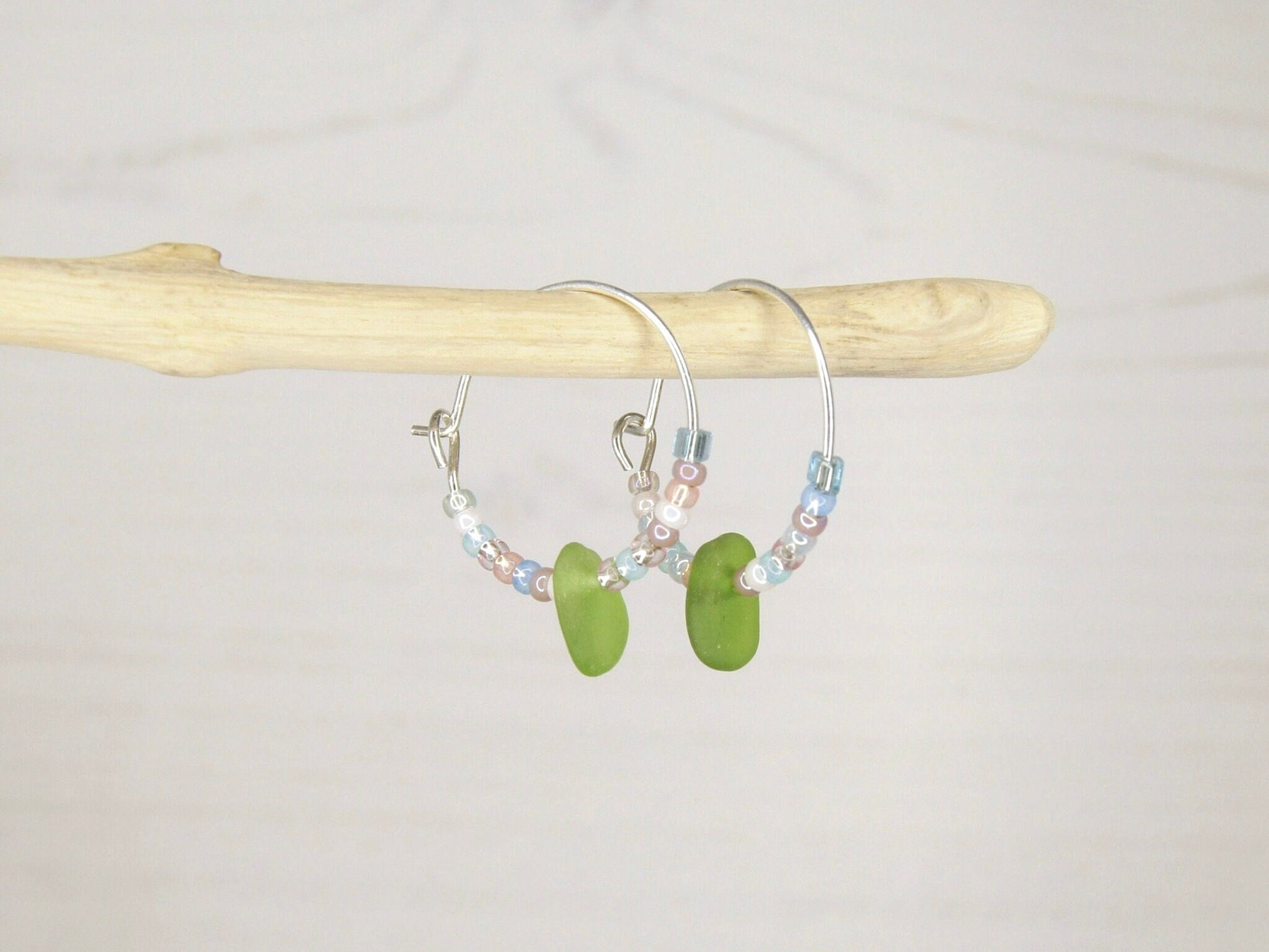 Hoop earrings with pastel seed beads and green sea glass hanging on driftwood against a light background