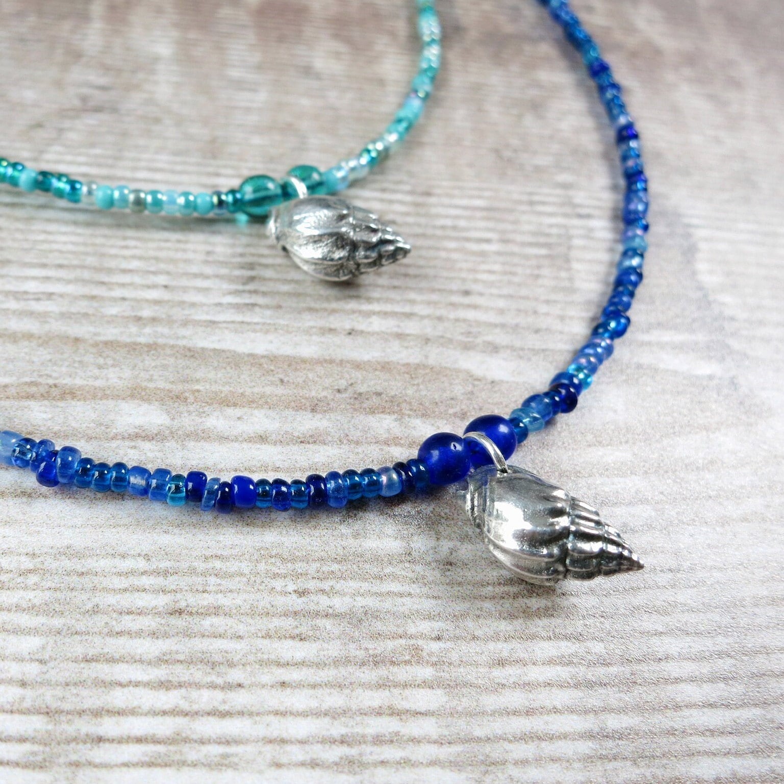Two seed bead necklaces with a silver shell pendant laid on a textured surface