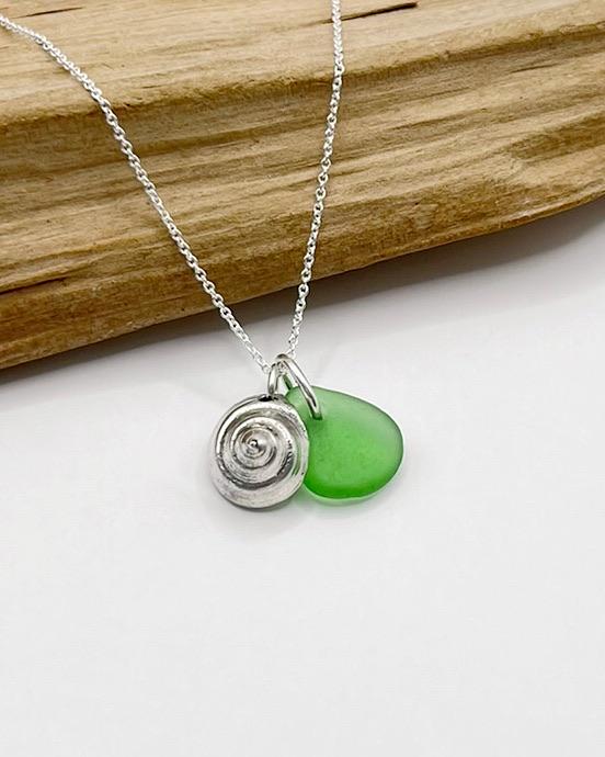 Necklace with a silver shell and green sea glass laid across driftwood against a white background