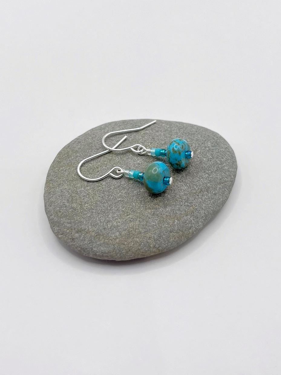 Turquoise beaded earrings laid on a grey pebble with a white background