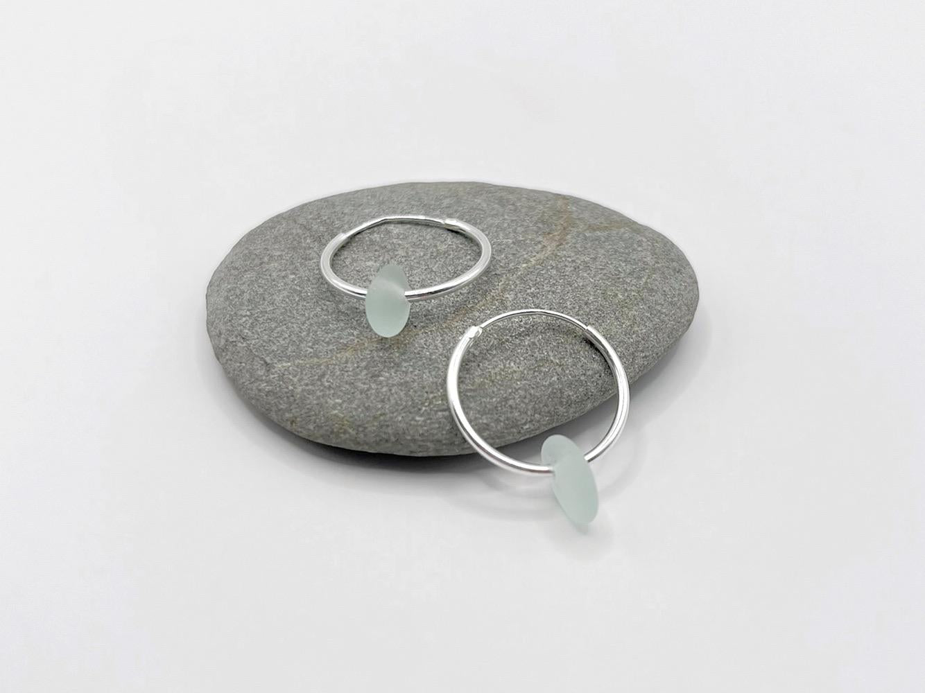 Silver hoop earrings with sea foam coloured sea glass laid on a grey pebble against a white background