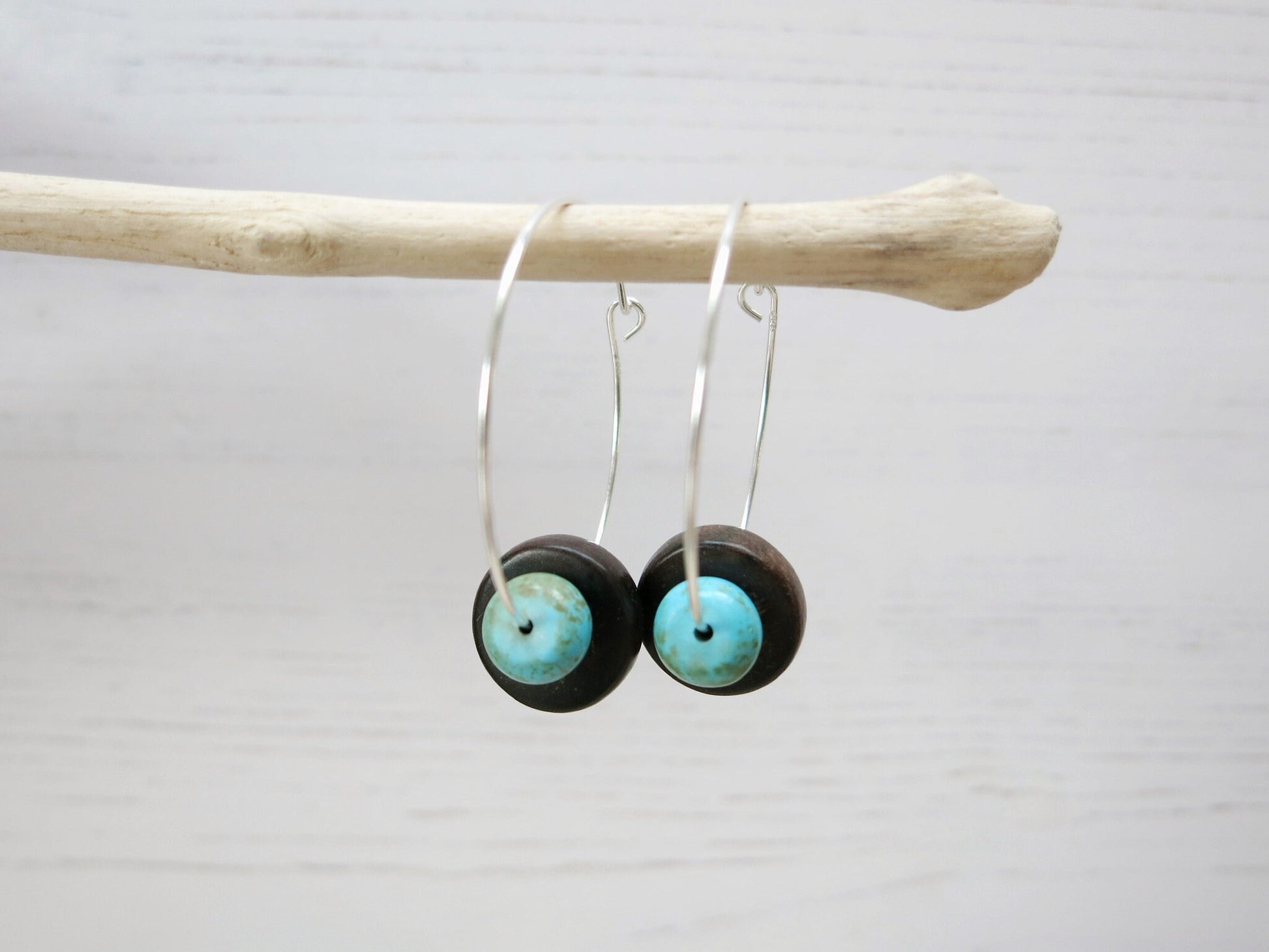 Close up of silver hoop earrings with turquoise and wooden beads hanging on driftwood against a light background