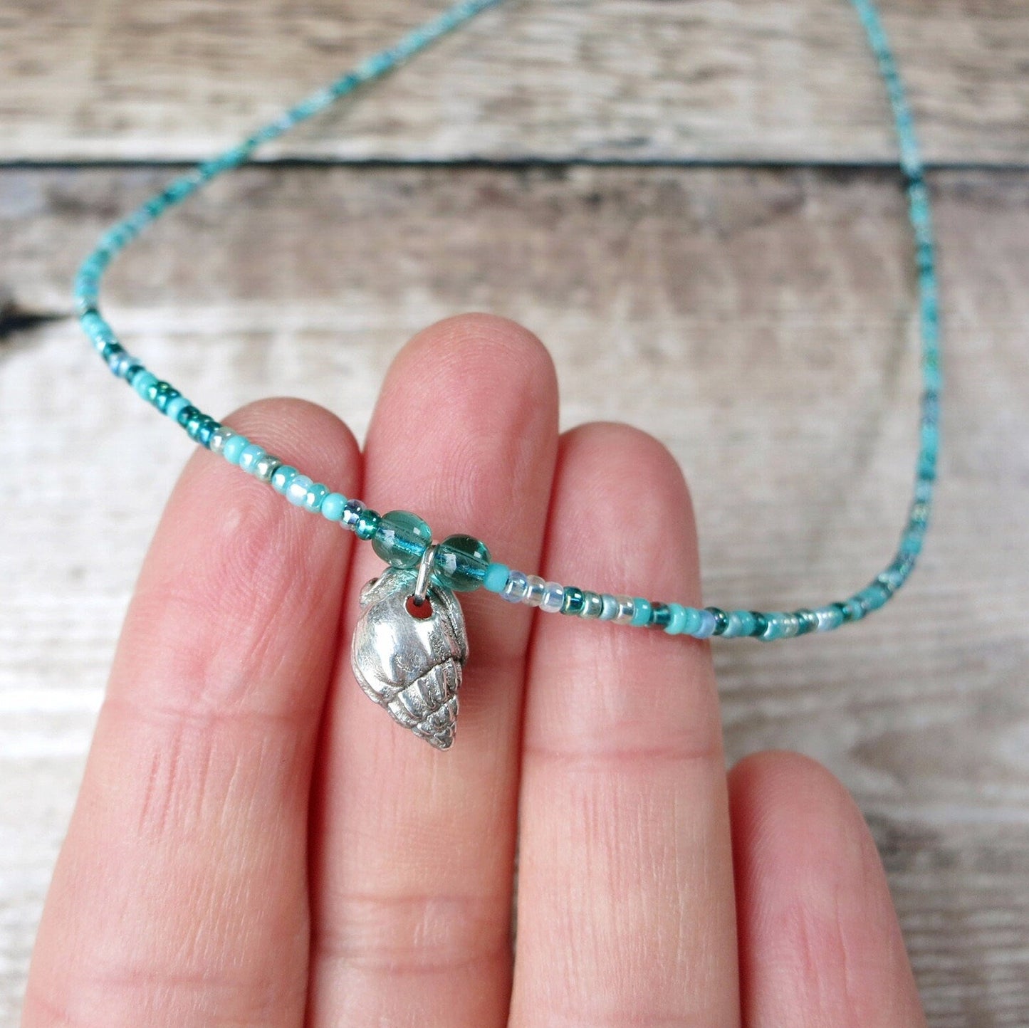 Turquoise seed bead necklace with a silver shell charm held between fingers over a wooden surface