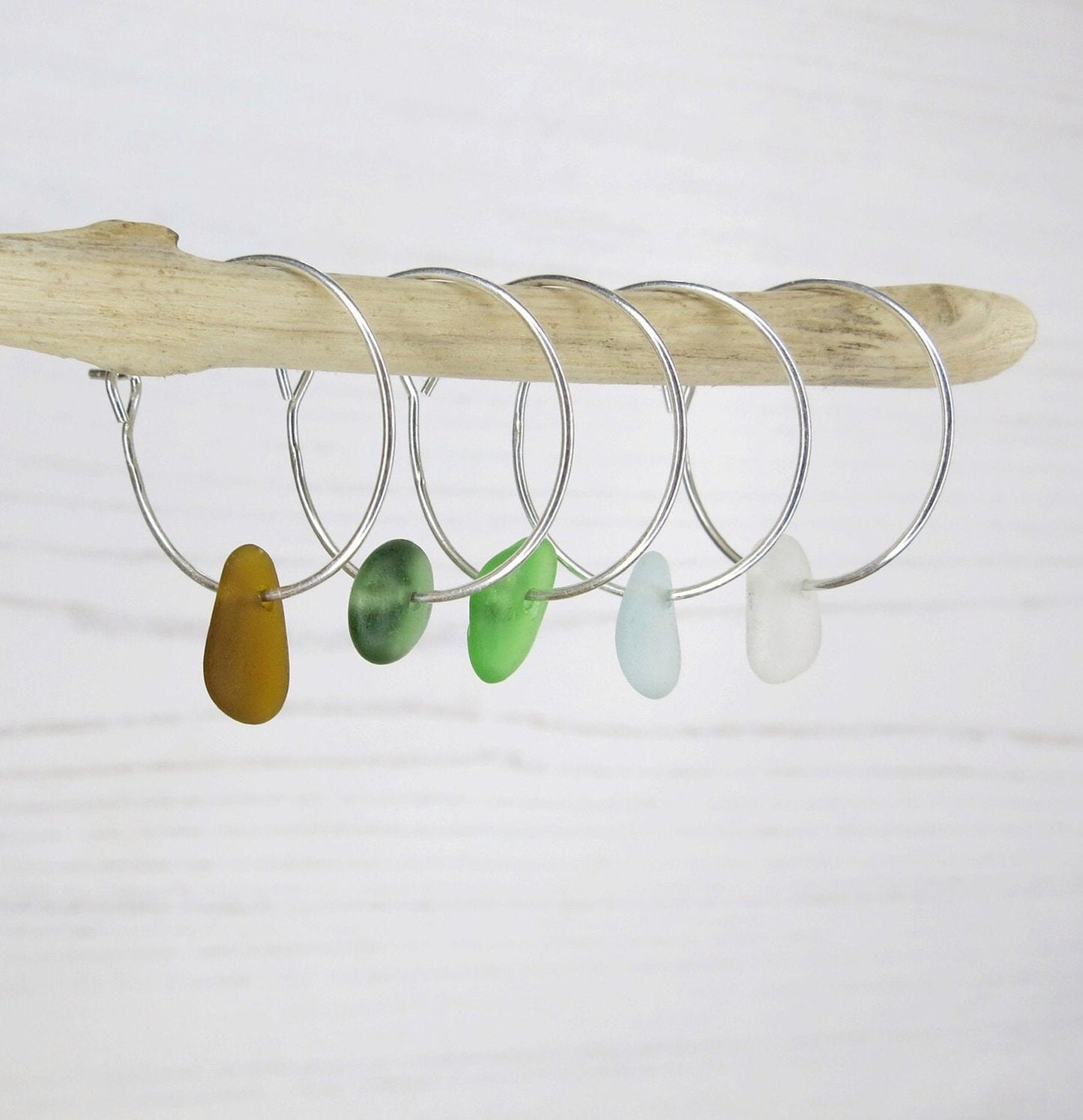 Silver hoop earrings with colorful sea glass hanging on a driftwood against a white background