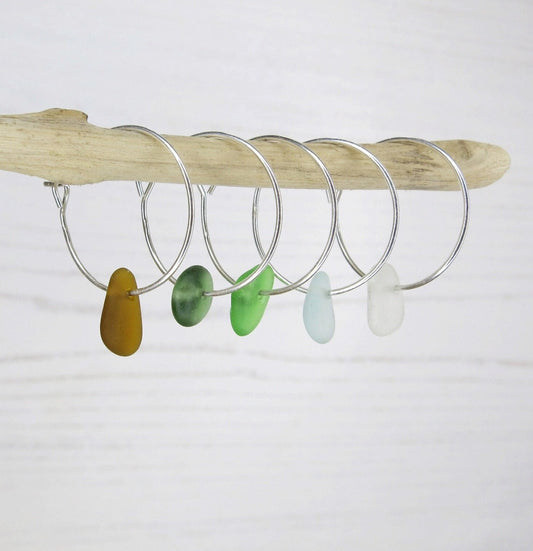 Silver hoop earrings with colorful sea glass hanging on a driftwood against a white background