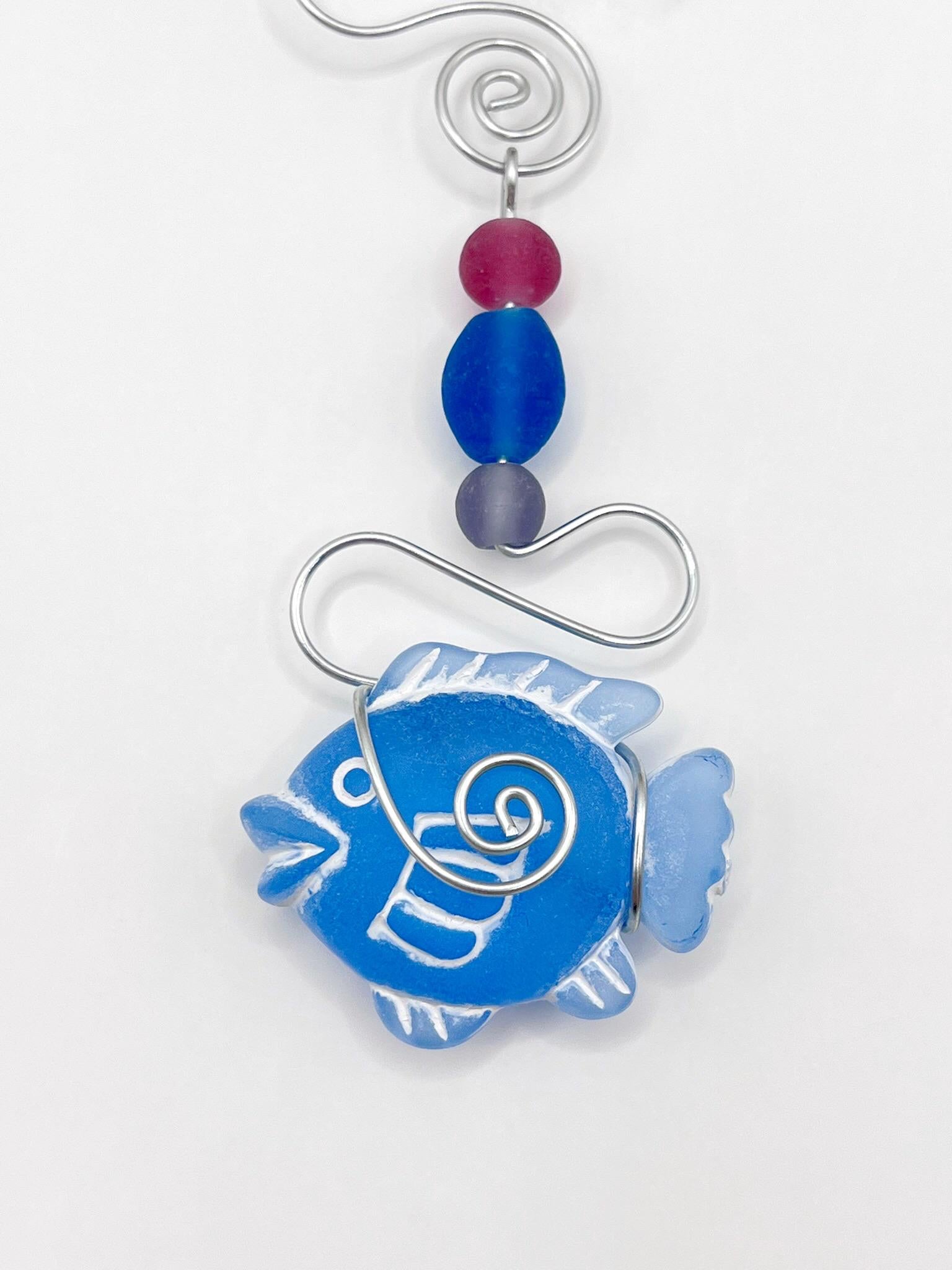 Close up of decorative hanging ornament with blue fish design, wire and colourful beads, laid  on a white background