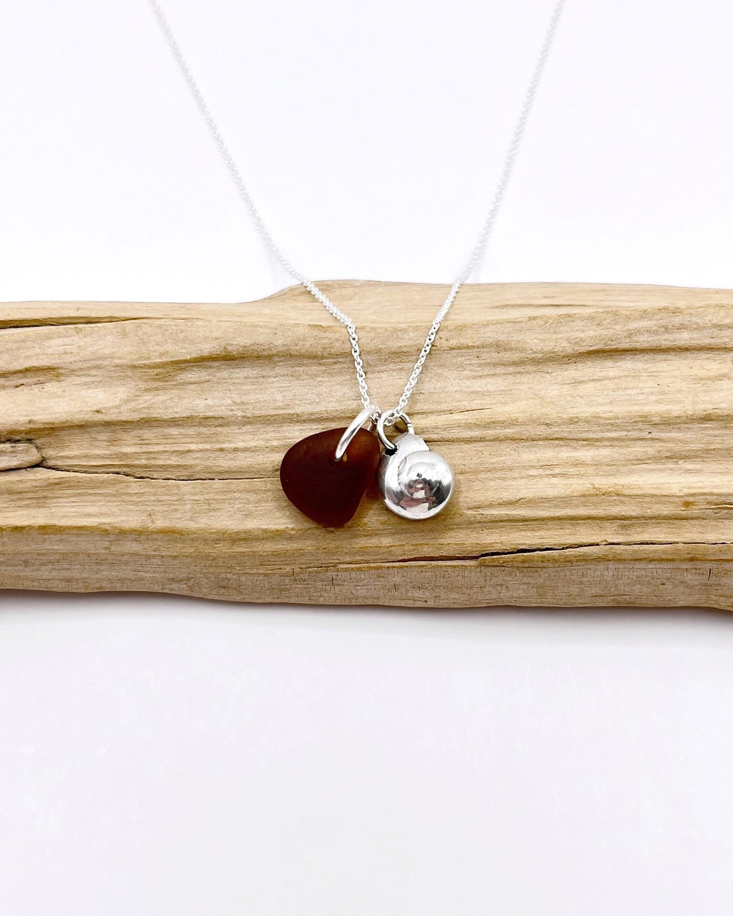Necklace with burnt orange sea glass and a silver shell pendant, laid on driftwood against a white surface