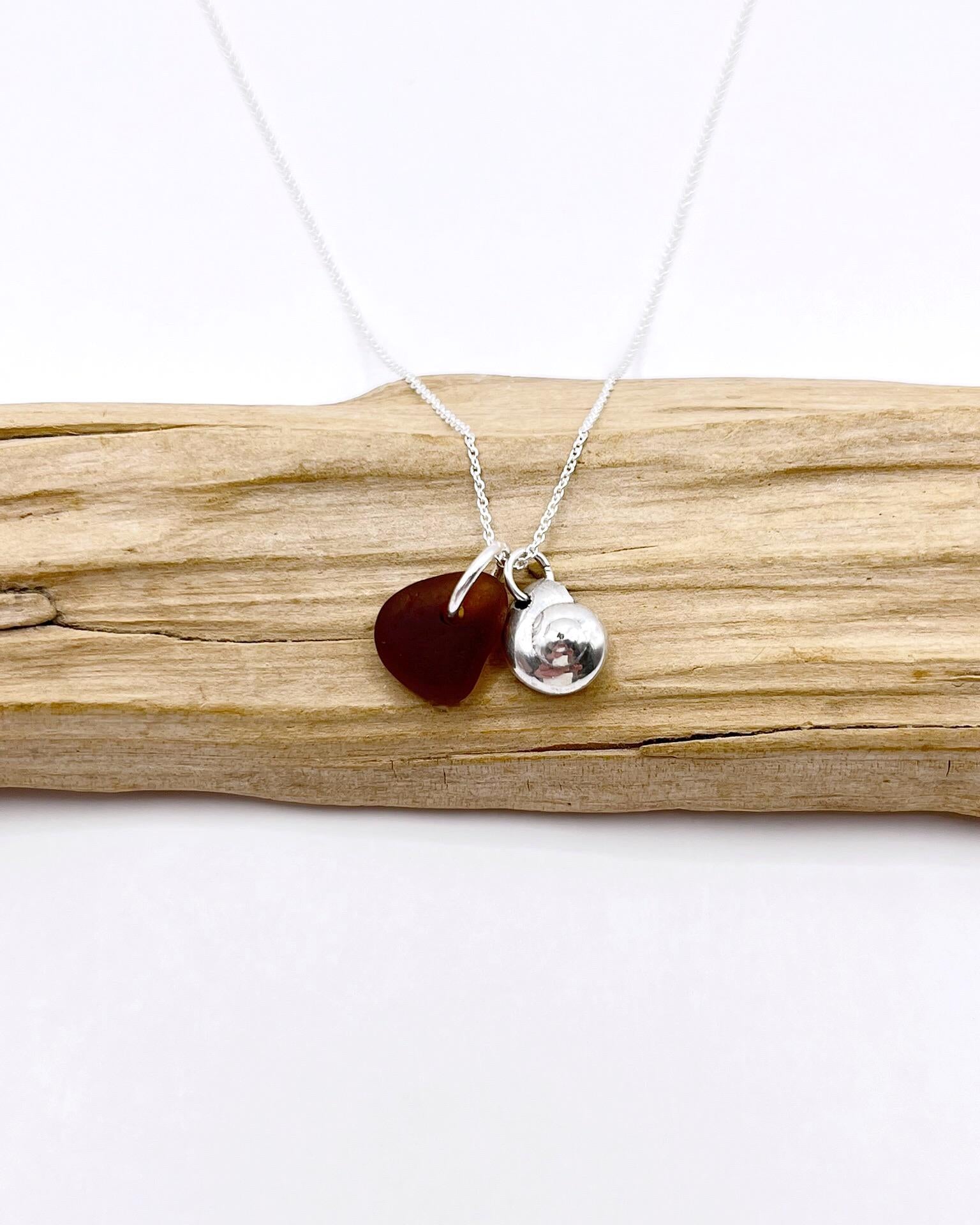 Necklace with burnt orange sea glass and a silver shell pendant, laid on driftwood against a white surface