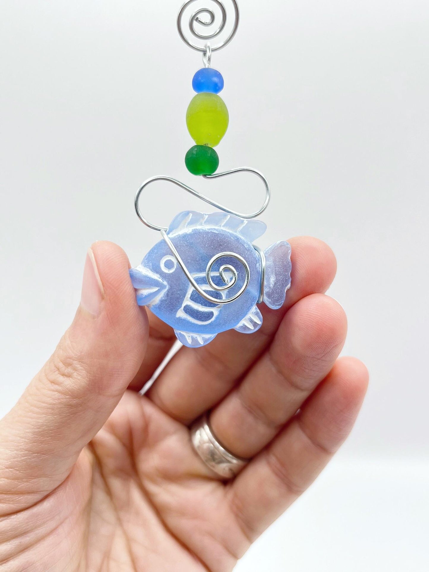 Decorative hanging with blue fish, wire and coloruful beads held in a hand against a white background