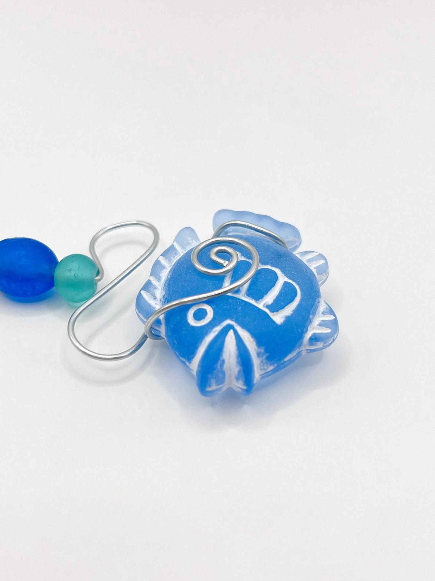 Close up of decorative ornament with blue fish design, wire and colourful beads on a white background